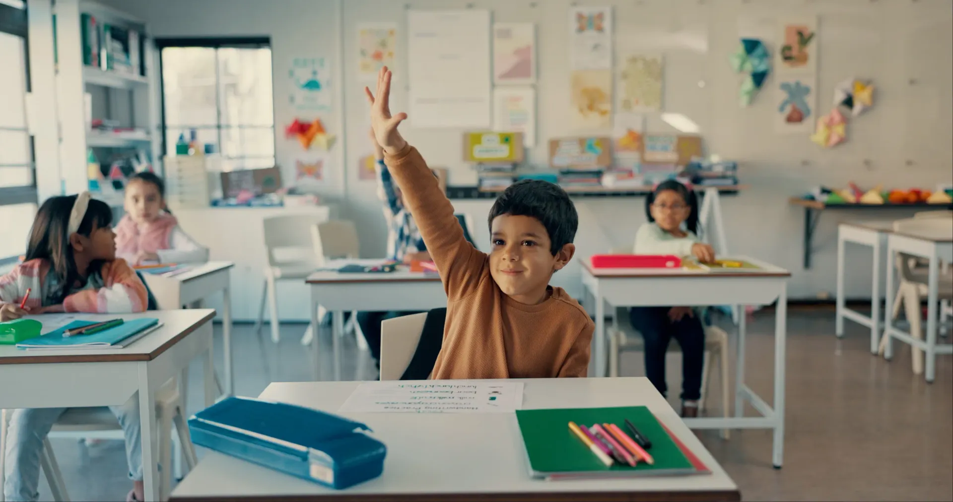 Un niño levanta la mano en un aula, con cuadernos y lápices sobre los pupitres en primer plano.