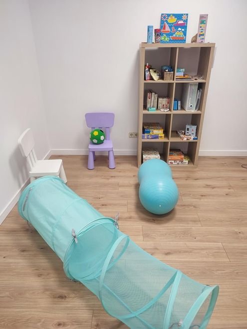 Sala de juegos infantil con una silla blanca, un túnel azul, una pelota de equilibrio, un taburete morado y una estantería sobre un suelo de madera.