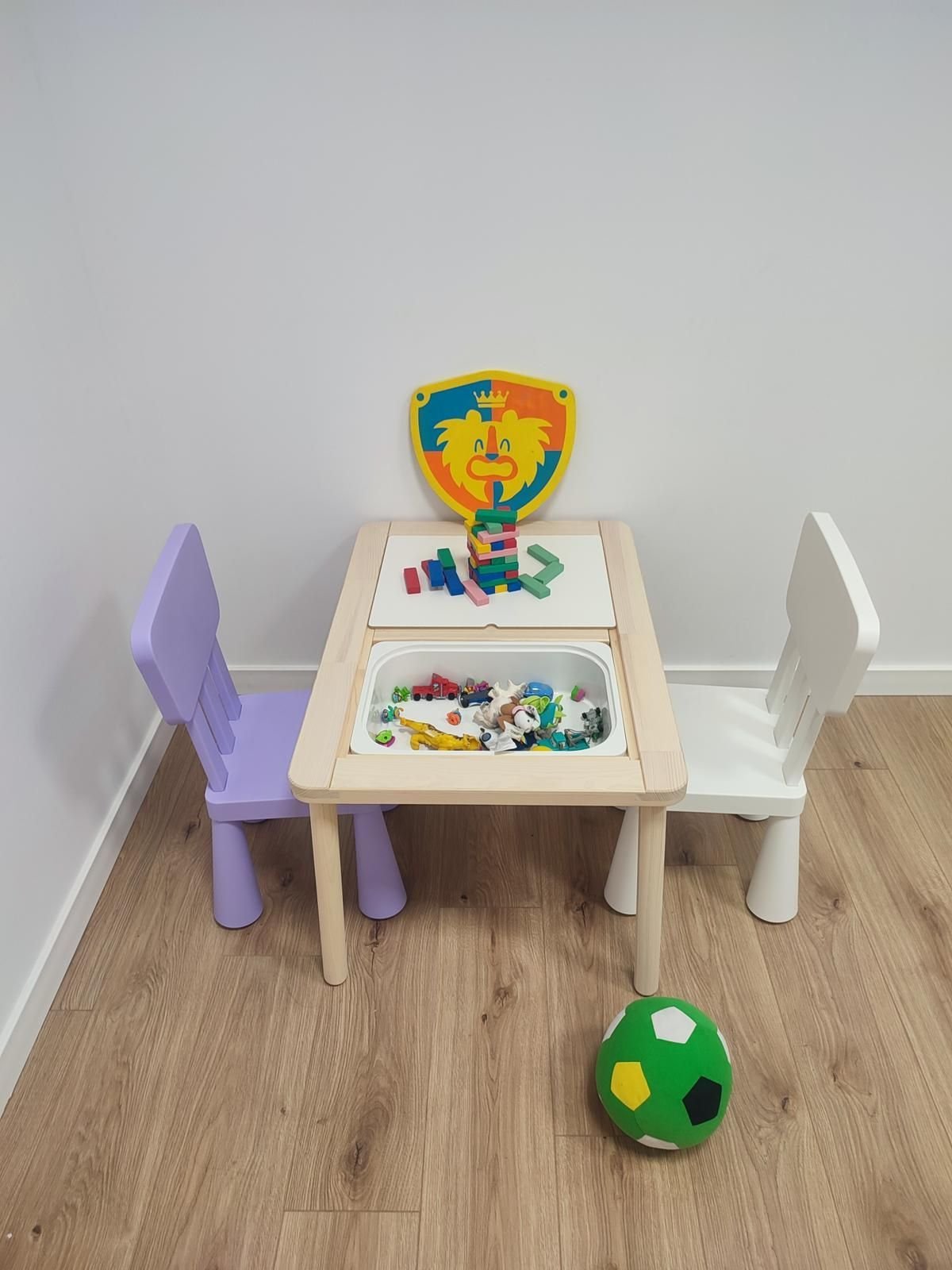 Mesita de juguete con dos sillas, un escudo encima y una pelota verde sobre un suelo de madera.