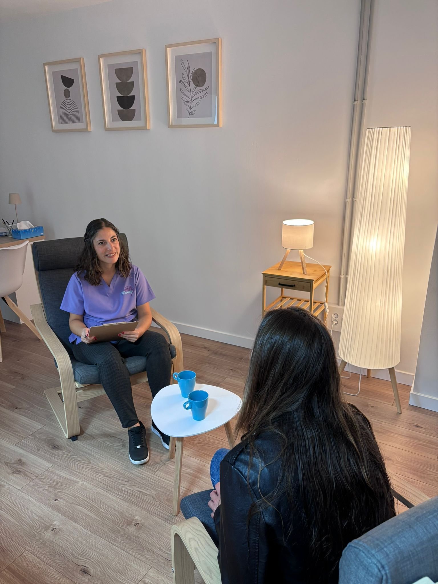 Dos personas conversando en una acogedora sala de terapia con sillas, una mesita, cuadros en la pared y una lámpara de pie.