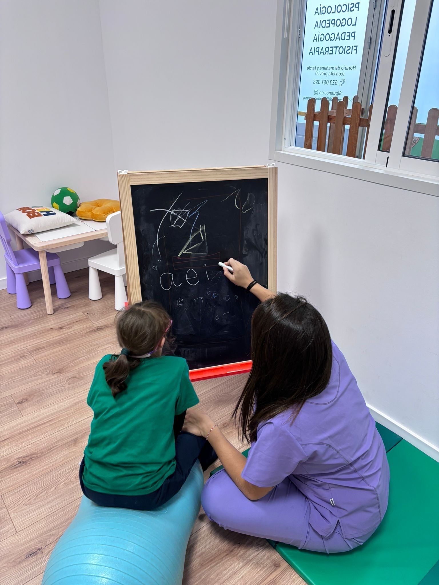 Dos niños están sentados sobre cojines en el suelo, dibujando en una pizarra en una sala de juegos.
