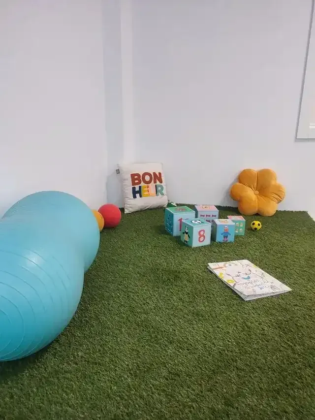 Zona de juegos con césped verde, un gran túnel azul, pelotas pequeñas, bloques de juguete y una silla naranja contra una pared blanca.